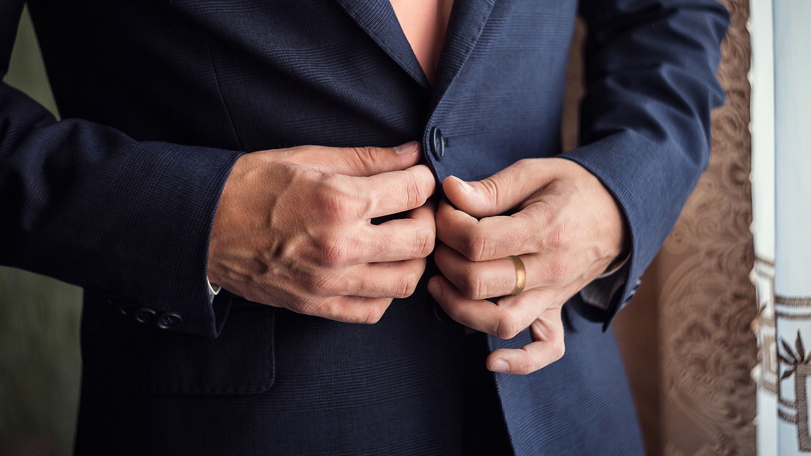 The man in the blue suit surgically wearing a yellow gold wedding ring is fastening his buttons